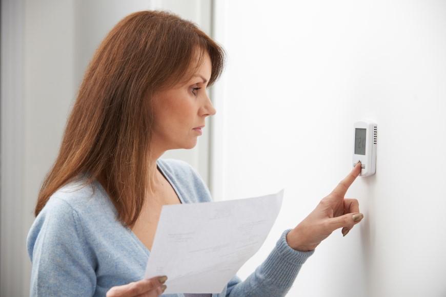 Woman with red, shoulder-length hair and wearing a light blue sweater. She is adjusting thermostat with her left index finger and holding a piece of paper in her right hand.