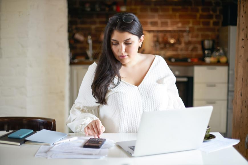 Younger woman at table using calculator and laptop