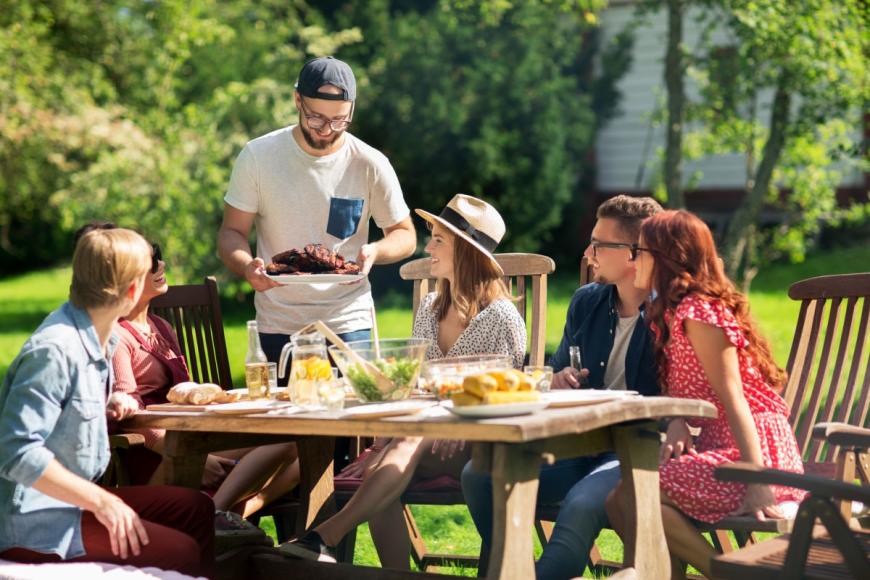 Young man serving plate of food to other young adults seated around table outside during the summer