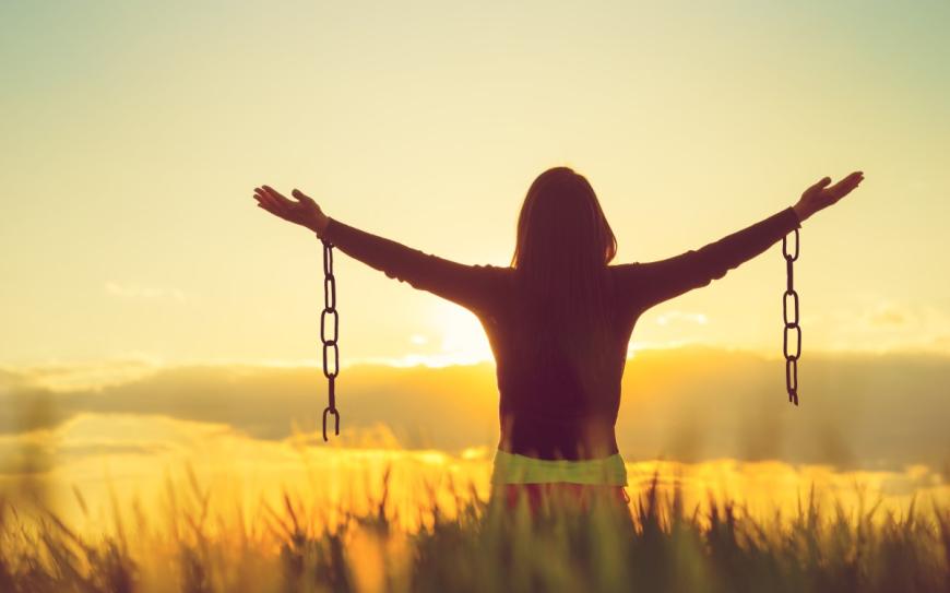 Woman facing sunset, arms extended widely to the sides. Each arm has a broken chain
