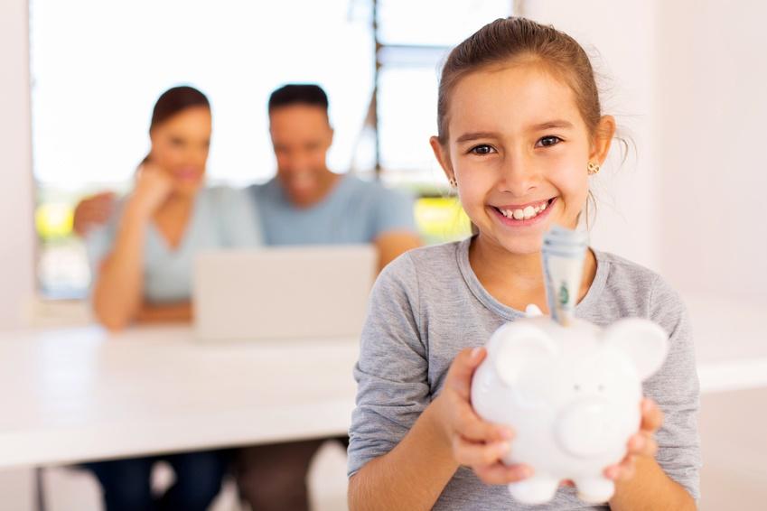 Young girl holding piggy bank and smiling. A couple in the background looking at information their laptop.