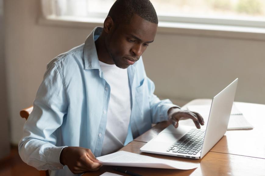 Man typing at a laptop while looking at paperwork