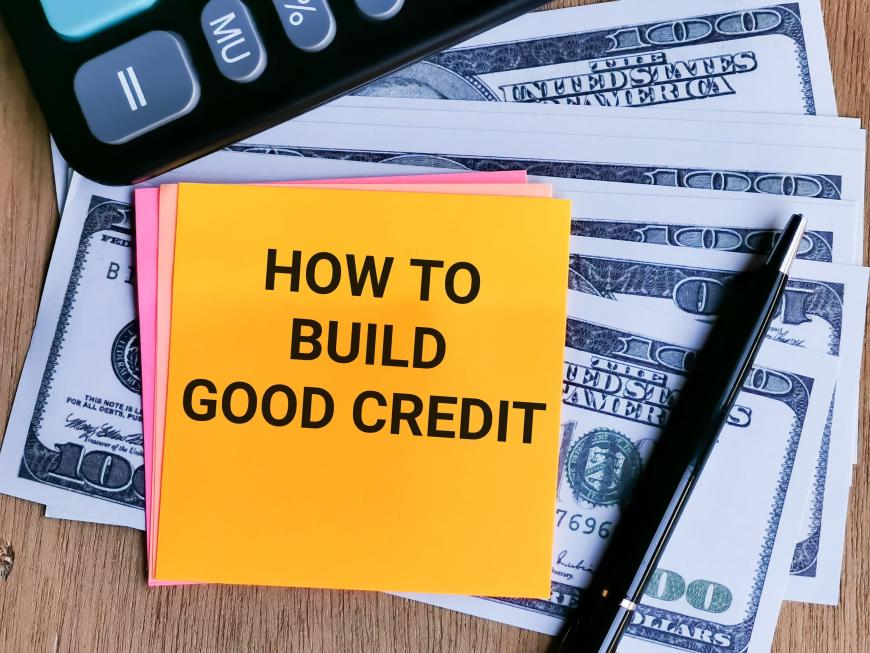 Post-it note reading "How to Build Good Credit." Post-it note resting on dollar bills and lying next to pen and keyboard
