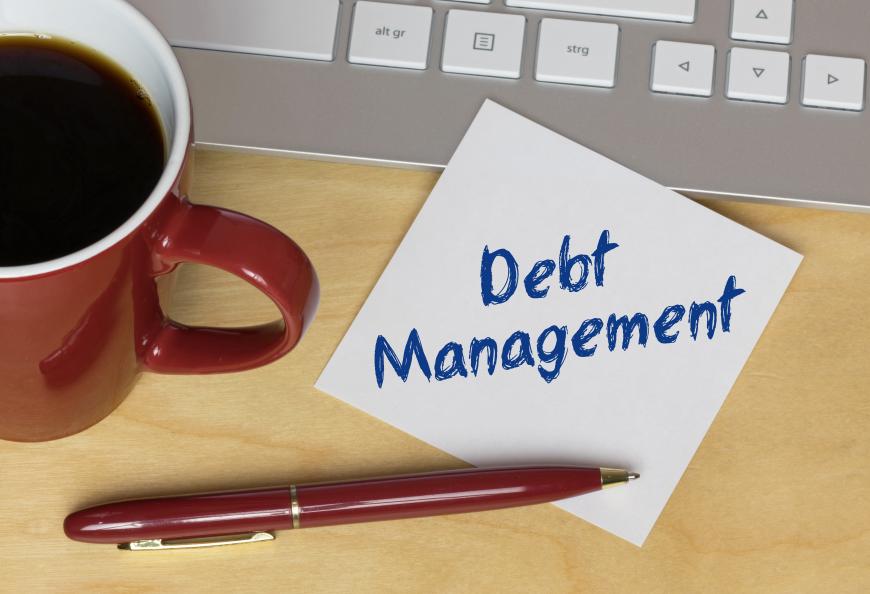 Post it that says Debt Management and resting on desk. Next to note are a cup of coffee, a computer keyboard, and a red pen.