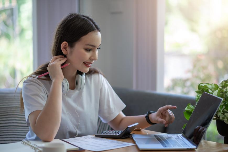 Young adult woman wearing headphones around her neck, sitting in front of her laptop with a calculator and notebook next to her. She is smiling and pointing at the laptop screen as she looks at it.