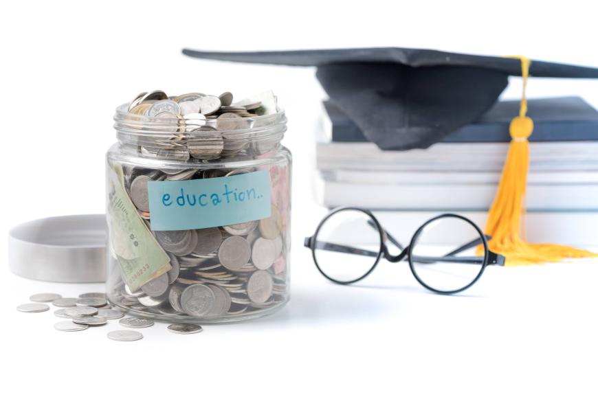 Jar filled with coins and labeled Education. To right of jar are a small stack of books with a graduation cap on top of them.