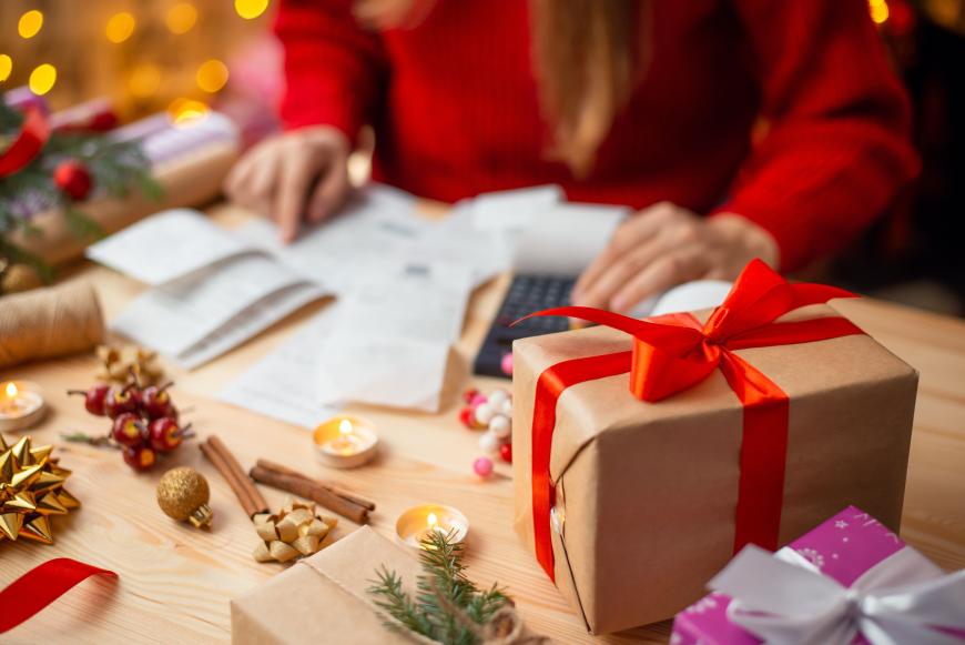Wrapped gifts on table and holiday ornaments next to them. Behind that is a person calculating gift expenses.