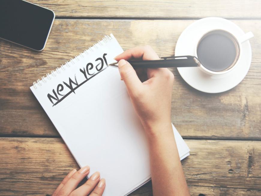 Person writing "New Year" on a sheet of paper. A cup of coffee is on the table to the right of person; a smart phone lies on the table above the sheet of paper.