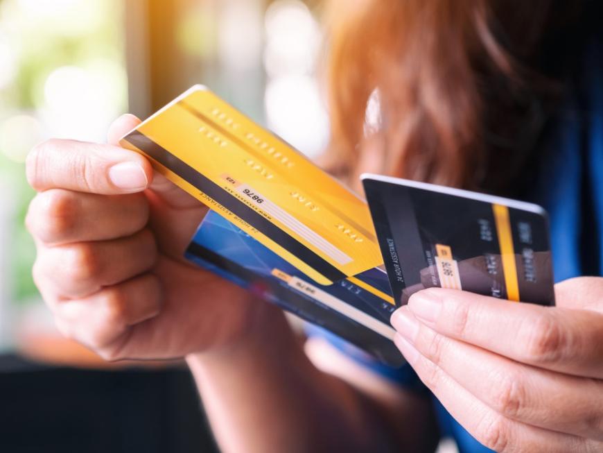 Person holding three credit cards (one black, one yellow, and one blue) with the left hand and pulling the yellow card from the pile with the right hand.