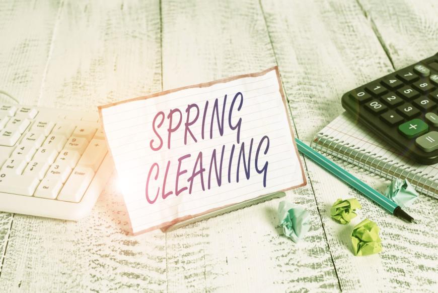 A note with the words SPRING CLEANING. To the left of the note is a white keyboard. To the right of the note are a pen; a black calculator; a spiral notebook; and small, crumpled post-it notes of different colors.