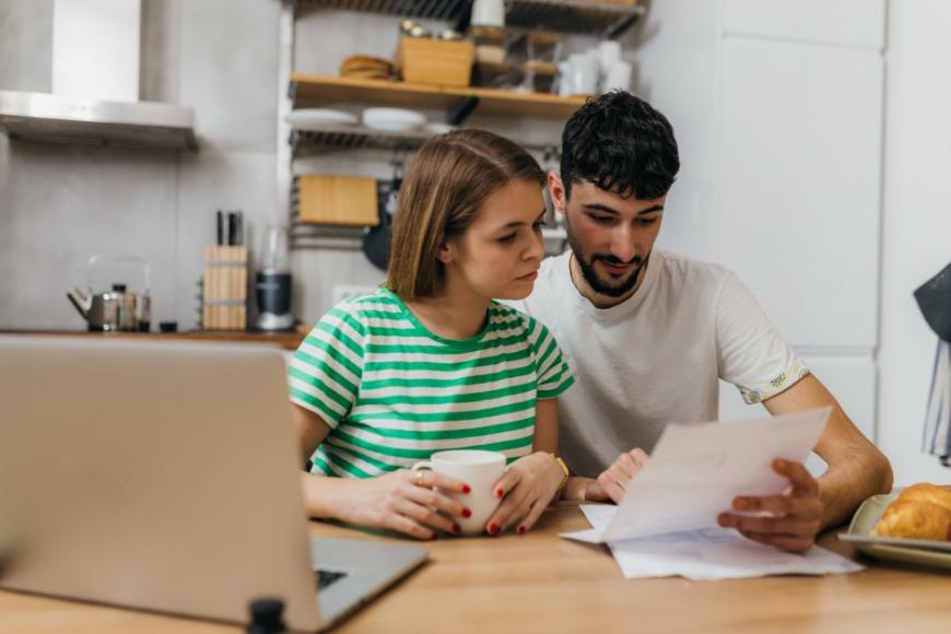 A woman and man sitting at a table and looking at documents together. The man is pointing at something in the document. A laptop computer is on the table to the left.