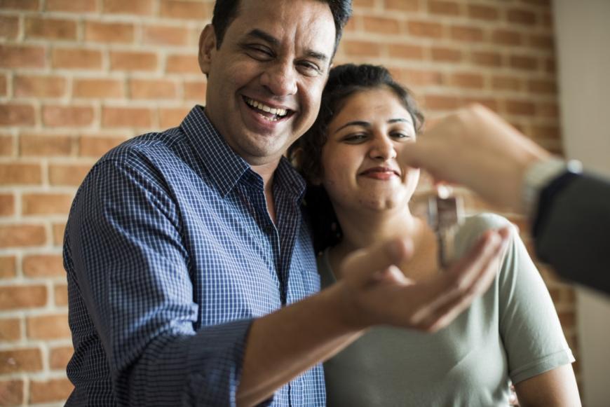 Couple with arms around each other, smiling. Person off camera puts a key in the man's hand.