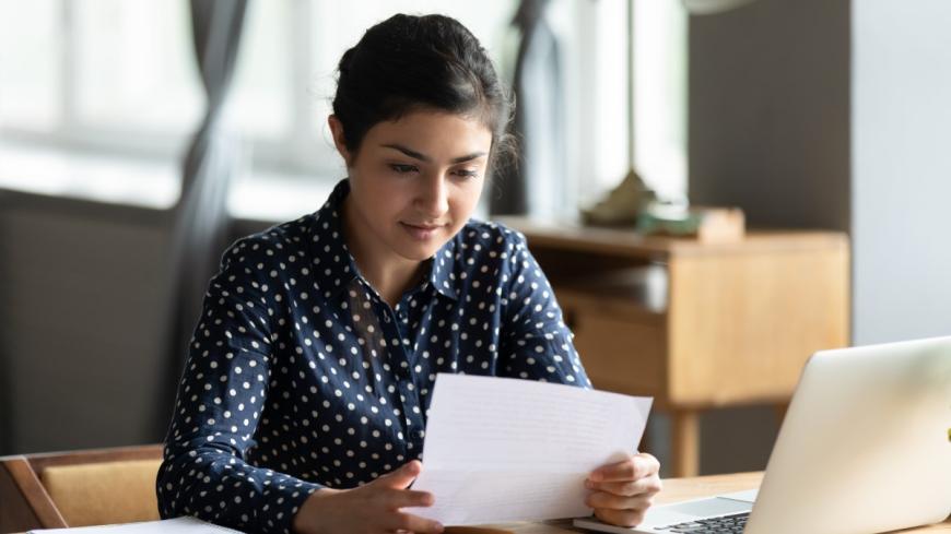 Young woman of south Asian descent, sitting at a table, holding a document a document, and reading it intently. On the table in front of her is a laptop.
