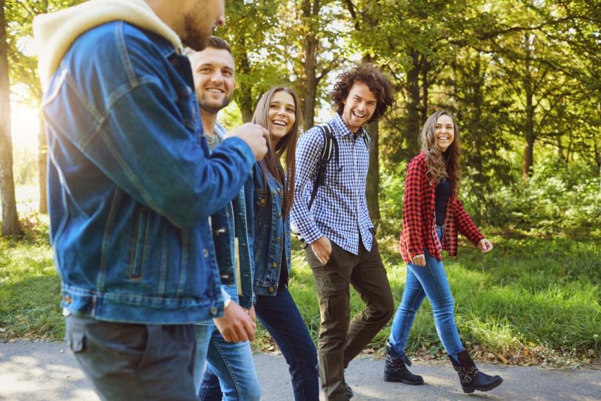 Three adult men and two adult women walking and laughing together along a path in a wooded area.
