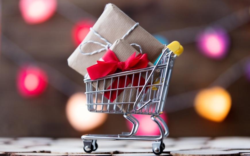 Metal shopping cart with two wrapped presents in its basket. Behind the cart are blurry images of multi-colored lights.