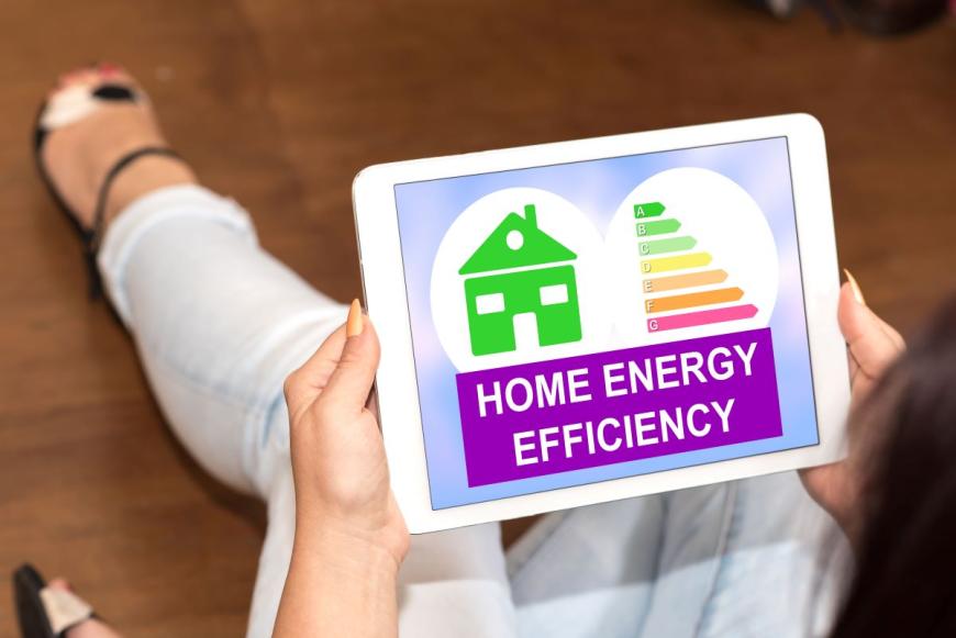 Woman sitting on the floor with her knees brought up toward her body and reading a computer tablet that is resting on her knees. The tablet screen displays a green graphic of a house and the text "Home Energy Efficiency."