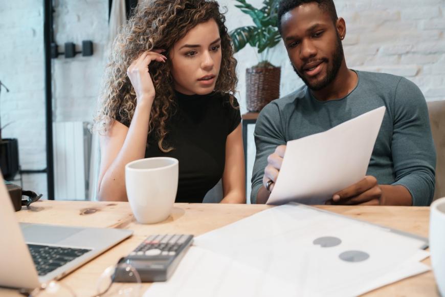 A couple sitting at table and looking over a document. The man sits on the right and points to something in the document. The woman is on the left and looks at what the man is pointing at. On the table in front of them are a coffee mug, a calculator, more documents, and a laptop computer.