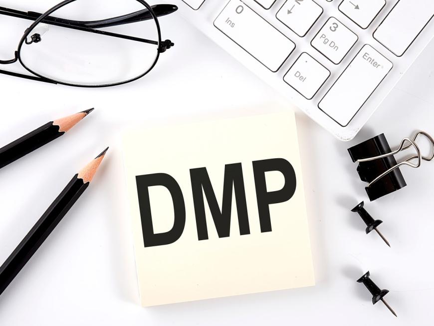 Post-it note with letter DMP in bold black type. Surrounding the note are two tacks, one black binder clip, two black pencils, a pair of eyeglasses, and a computer keyboard.