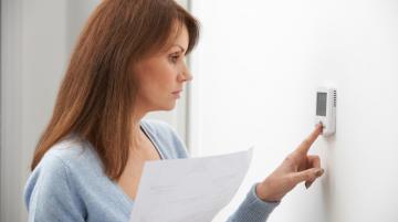 Woman with red, shoulder-length hair and wearing a light blue sweater.  She is adjusting thermostat with her left index finger and holding a piece of paper in her right hand.