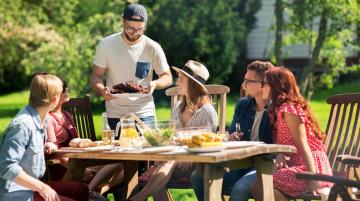 Young man serving plate of food to other young adults seated around table outside during the summer