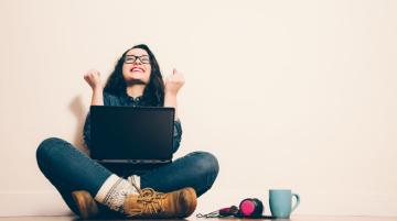 Young woman sittling on the floor with laptop in her lap, clenching her fists in a victory pose