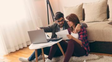 Couple sitting on floor in front of couch, using a laptop and looking at papers