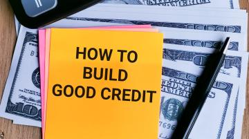 Post-it note reading "How to Build Good Credit."  Post-it note resting on dollar bills and lying next to pen and keyboard