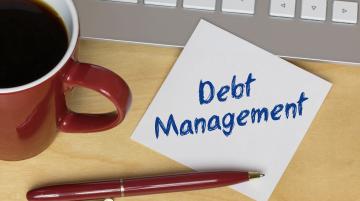 Post it that says Debt Management and resting on desk.  Next to note are a cup of coffee, a computer keyboard, and a red pen.