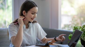 Young adult woman wearing headphones around her neck, sitting in front of her laptop with a calculator and notebook next to her.  She is smiling and pointing at the laptop screen as she looks at it.
