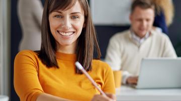 Adult woman sitting at desk, writing on papers, and smiling for the camera. Man sitting behind her and works on a laptop.  Two women stand behind man in front of a whileboard.