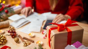 Wrapped gifts on table and holiday ornaments next to them. Behind that is a person calculating gift expenses.