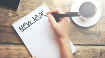 Person writing "New Year" on a sheet of paper.  A cup of coffee is on the table to the right of person; a smart phone lies on the table above the sheet of paper.