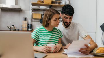 A woman and man sitting at a table and looking at documents together.  The man is pointing at something in the document.  A laptop computer is on the table to the left.