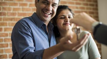 Couple with arms around each other, smiling.  Person off camera puts a key in the man's hand.