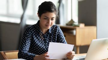 Young woman of south Asian descent, sitting at a table, holding a document a document, and reading it intently.  On the table in front of her is a laptop.