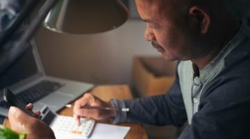 Person of color sits at a desk, making calculations on calculator with the right hand and holds a cell phone with the left hand.  A laptop lies on the table behind the calculator.