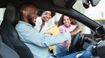 Happy Black man sitting in the driver's seat of a car and holding the steering wheel.  Black woman and black child smile at him from outside the driver's side of the car.