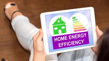 Woman sitting on the floor with her knees brought up toward her body and reading a computer tablet that is resting on her knees.  The tablet screen displays a green graphic of a house and the text "Home Energy Efficiency."