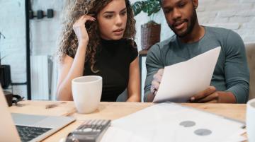 A couple sitting at table and looking over a document.  The man sits on the right and points to something in the document.  The woman is on the left and looks at what the man is pointing at.  On the table in front of them are a coffee mug, a calculator, more documents, and a laptop computer.