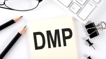 Post-it note with letter DMP in bold black type.  Surrounding the note are two tacks, one black binder clip, two black pencils, a pair of eyeglasses, and a computer keyboard.