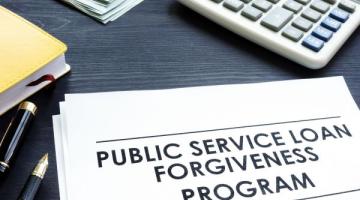 A stack of papers on a desk; the top sheet in the stack reads Public Service Loan Forgiveness Program in bold, black type and all capital letters.  Also on the table are three pens, a notebook, a stack of money with a $10 bill on top, and a calculator.