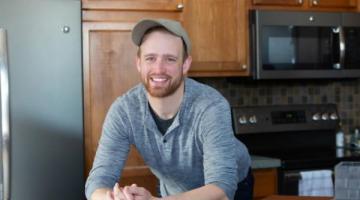 Man wearing tan cap and long sleeved blue patterned shirt.  He smiles for the camera and leans on his elbows on a kitchen counter.  He has red beard and mustache.