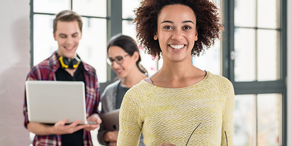 Woman holding glasses and smiling, behind her, a two people look at a laptop.