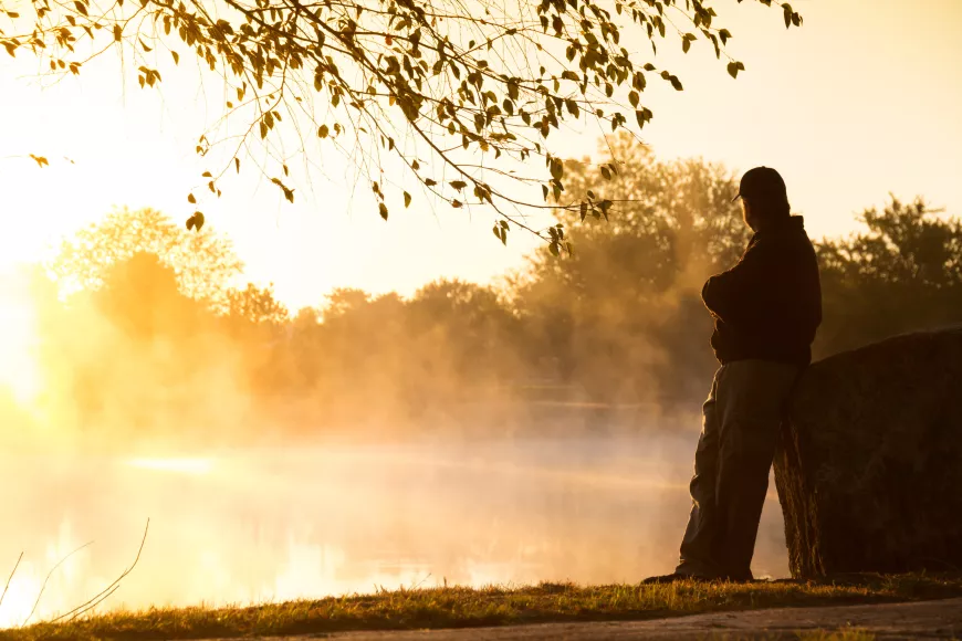 Man leaning against a rock at the edge of a body of water, looking at the sunrise.