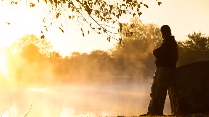 Man leaning against a rock at the edge of a body of water, looking at the sunrise.
